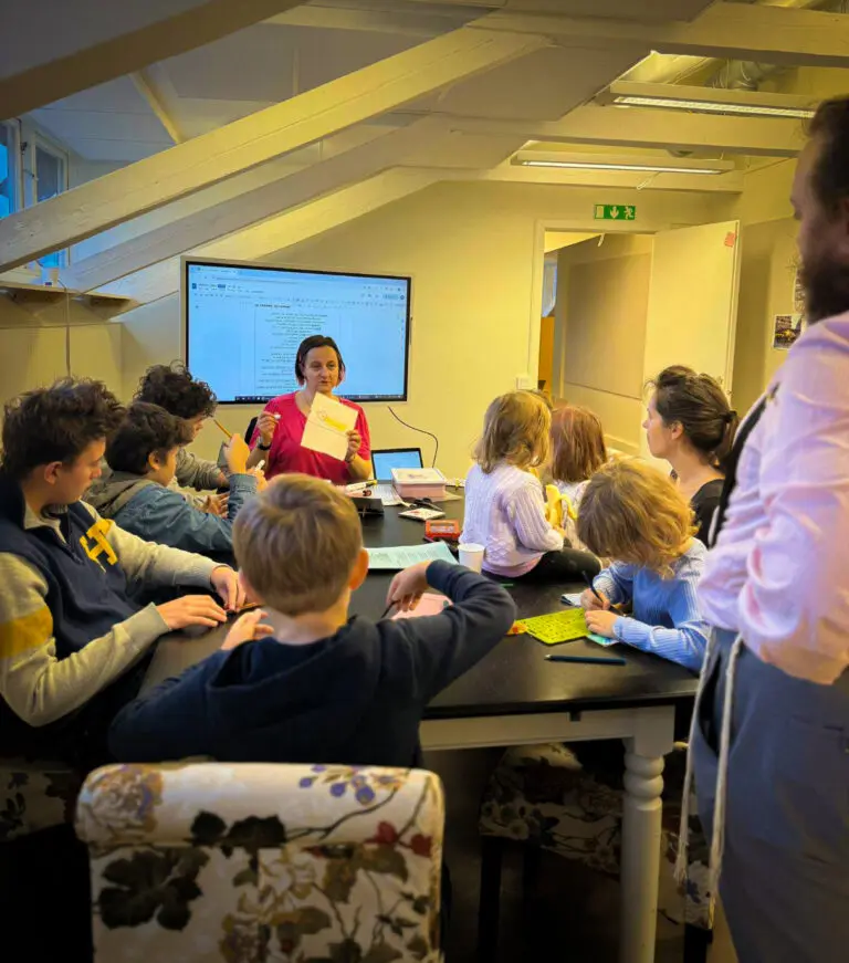 A group of children and a young teenager, together with two parents around a table. The teacher Katka is showing a piece of paper in front of a screen.